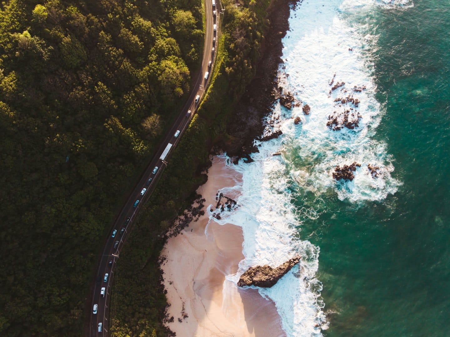Cars travel along a coastal road beside the sandy beach.